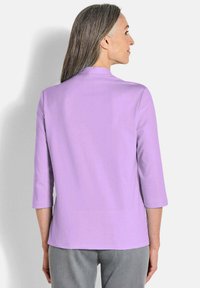 Lila top met driekwartmouwen, rechte snit, zachte textuur en ronde hals, gecombineerd met grijze broek, van achteren gezien.