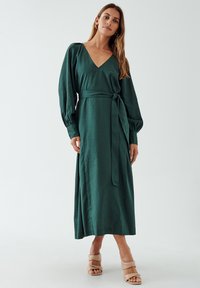 Long green dress with a deep V-neck, cinched waist, and long sleeves. Made from a textured fabric, featuring side ties and pleats.