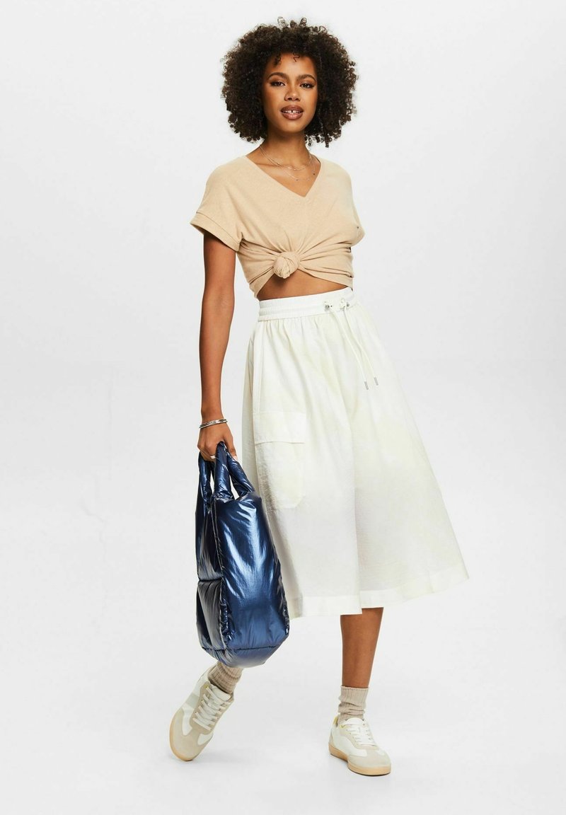 Beige Cropped-Top mit Knoten-Detail, weiße Midirock, hält eine glänzende blaue Tasche, cremefarbene Sneakers, leicht strukturierter Stoff.