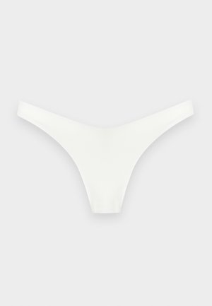 Žena stojaca bosá v bielych bikinách so štvorcovým výstrihom, s jednou rukou na bokoch na jednoduchom svetlom pozadí.