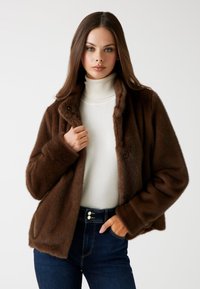 Giacca in pelliccia sintetica marrone con colletto, bottoni frontali e maniche lunghe, indossata sopra un dolcevita bianco e jeans scuri.