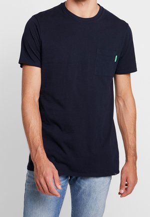 Mężczyzna noszący prosty czarny T-shirt z krótkim rękawem i kieszenią na piersi oraz jasnoniebieskie dżinsy, stojący na jednolitym tle.