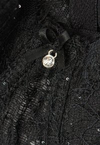 Zwarte kant kledingstuk met een klein strikje en een zilveren charm met een heldere steen. De stof heeft een gestructureerd patroon met glinsterende pailletten.