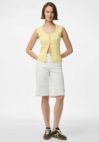Gul ribbad ärmlös topp med v-ringning och scallop-kant, kombinerad med vita knälånga shorts och bruna sneakers.