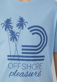 Hellblaues Baumwoll-T-Shirt mit blauen Palmen-Grafiken und geschwungenen Linien. Der Text lautet "OFFSHORE PLEASURES" in einer kontrastierenden Schriftart.