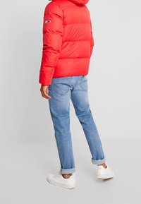 Rød pufferjakke med horisontal polstring, hætte og logo på ærmet, parret med lyseblå jeans og hvide sneakers.