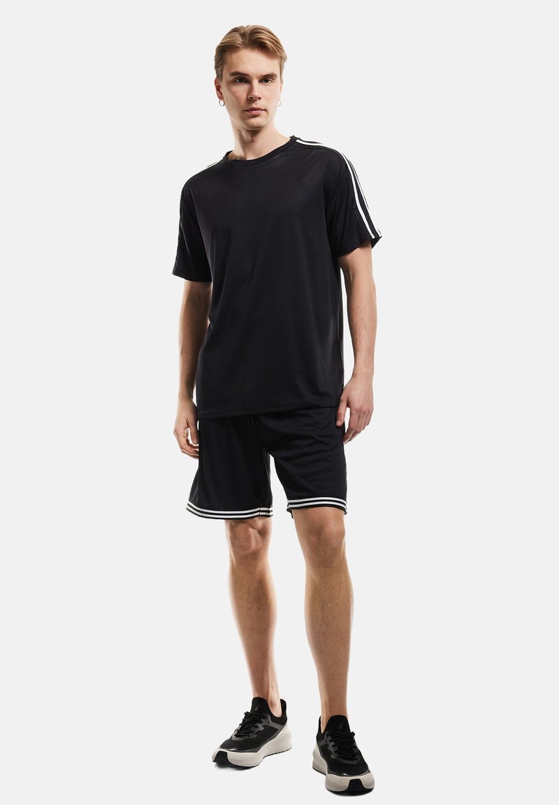 T-shirt de sport noir avec des rayures blanches sur les manches, associé à un short noir avec des ourlets à rayures blanches, porté avec des baskets noires.