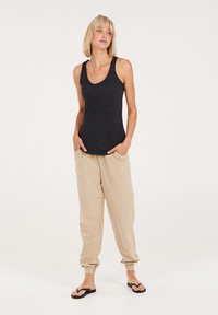 Schwarzes Tanktop mit breiten Trägern, kombiniert mit beigen, lockeren Jogginghosen, die elastische Bündchen und seitliche Taschen haben. Schwarze Sandalen.
