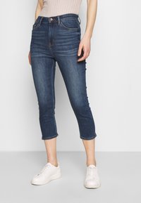 Jeans capri de mezclilla azul oscuro con una cintura alta, ajuste ceñido, bolsillos frontales y dobladillos enrollados, combinados con zapatillas blancas.
