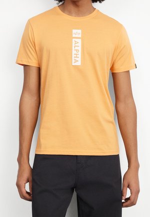Jeune personne portant un t-shirt orange à manches courtes avec un logo blanc vertical "ALPHA" et un petit emblème au centre, associé à un pantalon noir.