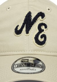 Căciulă bej ajustabilă, cu un logo brodat texturat în albastru închis "NE", având o etichetă rotundă neagră pe care scrie "New Era 9TWENTY."