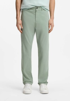 Uomo che indossa pantaloni casual verde chiaro e sneakers bianche, in piedi davanti a uno sfondo bianco semplice.