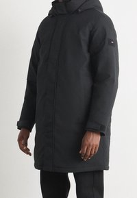 Svart vadderad jacka med huva, med en framdragkedja, justerbara ärmslut och ett logotyp-patch på vänster ärm. Slät, strukturerad tyg.