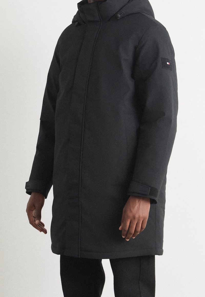 Svart vadderad jacka med huva, med en framdragkedja, justerbara ärmslut och ett logotyp-patch på vänster ärm. Slät, strukturerad tyg.