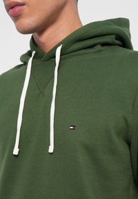 Zaļš džemperis ar mīkstu audumu, aprīkots ar baltu auklu un mazu logo sarkanā, baltā un zilā krāsā uz krūtīm.