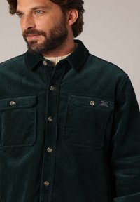 Grünes Cordhemd mit Knopfleiste, zwei Brusttaschen und strukturiertem Stoff. Verfügt über einen Kragen und braune Knöpfe.