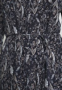 Granatowa sukienka z materiału w wzór paisley z wiązaniem w talii i długimi rękawami. Posiada skomplikowane wzory w odcieniach kremowego i jasnoniebieskiego.