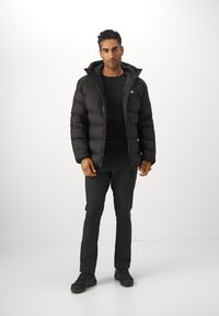 Doudoune noire avec capuche, présentant un design matelassé horizontal et une fermeture éclair. Portée sur un t-shirt à manches longues noir et un pantalon sombre.