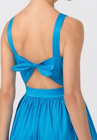 Blå klänning med utskärning i ryggen, breda axelband och en dekorativ rosett i midjan. Slät textur med en flödande kjol.