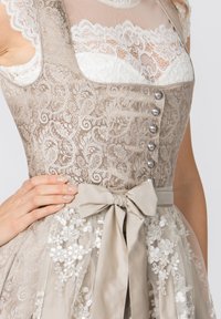 Dirndl-Kleid mit einem oberen Teil aus hellbeigem Spitzenmaterial mit Blumenmuster, silbernen Knöpfen und einer passenden Satin-Schleife in der Taille über einem transparenten Rock.