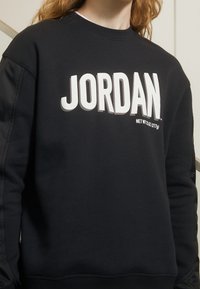 Sweat-shirt noir avec texte blanc "JORDAN", col rond côtelé et manches texturées. Tissu doux avec une coupe décontractée, détail de marque minimal.
