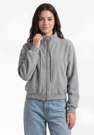 Ung kvinde med langt mørkt hår iført en grå fleecejakke med lynlås og lyseblå jeans, holder om jakke-kraven med den ene hånd.