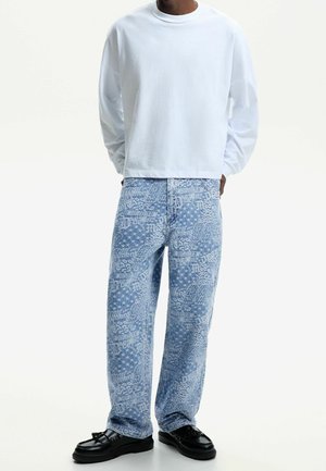 Osoba w gładkiej białej bluzce z długim rękawem, jasnoniebieskich szerokich spodniach dżinsowych z nadrukiem tekstu i gwiazdek oraz czarnych mokasynach z frędzlami.