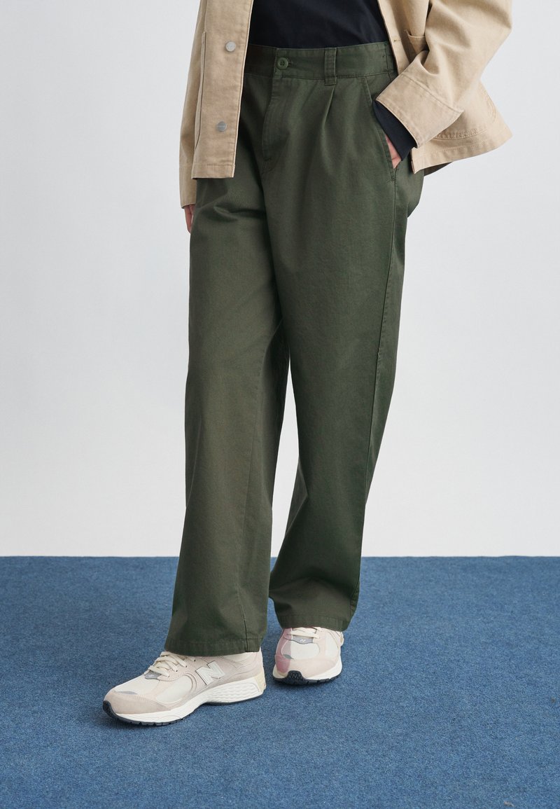 Άτομο που φοράει ελαιδί παντελόνι με φαρδιά μπάτη, μπεζ μπουφάν με τα μανίκια γυρισμένα, μαύρο πουκάμισο και μπεζ αθλητικά παπούτσια New Balance, στέκεται σε μπλε χαλί.