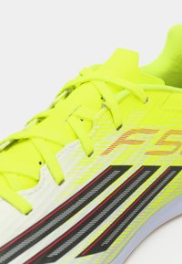 Φωτεινό κίτρινο αθλητικό παπούτσι με υφή στο επάνω μέρος, μαύρες και κόκκινες λεπτομέρειες, και επίπεδα κορδόνια. Διαθέτει κομψό, ρέον σχεδιασμό για απόδοση.