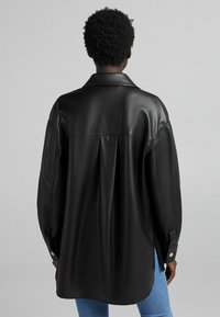 Black leather shirt jacket with a relaxed fit, featuring a back pleat, pointed collar, and buttoned cuffs. Smooth texture and minimal design.