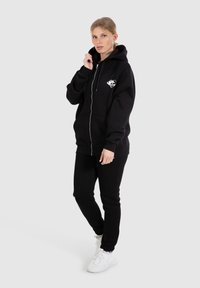 Hoodie noir zippé en polaire épaisse avec une poche avant et un graphique en forme de cœur. Associé à un pantalon de jogging noir et des baskets blanches.