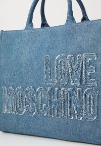 Denim tote bag med to håndtak på toppen, med brodert "LOVE MOSCHINO"-tekst i en slitt stil på en lys blå bakgrunn.