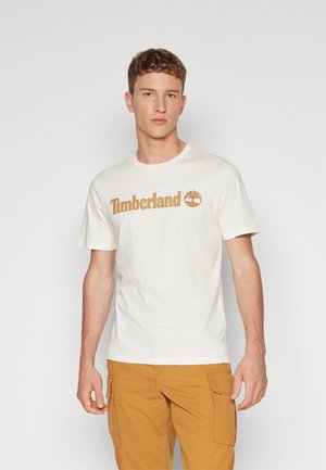 Cremefarbenes Baumwoll-T-Shirt mit braunem "Timberland"-Logo, Rundhalsausschnitt, kurzen Ärmeln, getragen mit beigen Cargo-Hosen.