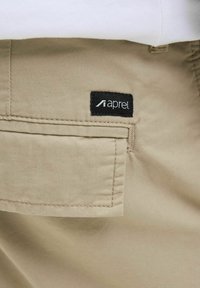 Beige fabric shorts with a side pocket, featuring a black label with the brand name "aprel." Smooth texture with neatly stitched edges.