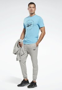 Camiseta azul de cuello redondo con un logo circular, pantalones de chándal grises con un logo en la cadera y zapatillas deportivas negras, con una sudadera gris colgada de un brazo.