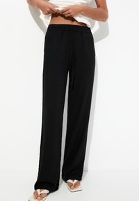 Pantalones negros, ligeros, con cordón y un ajuste holgado. La tela parece suave, con una textura lisa. Llevados con chanclas blancas.