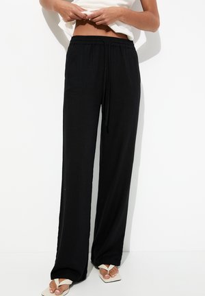 Pantalons noirs légers à cordon de serrage avec une coupe ample. Le tissu semble doux, avec une texture lisse. Portés avec des tongs blanches.