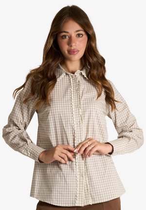 Donna con lunghi capelli castani che indossa una camicia beige a quadri con bottoni e rifiniture in pizzo, in piedi con le mani vicino ai bottoni, sfondo neutro.