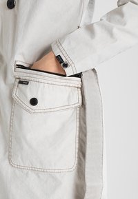 Giacca grigia chiara con tasca con zip e chiusura a bottoni. Presenta cuciture a contrasto e un'etichetta con logo sul polsino.