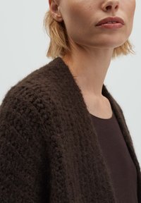 Donna con capelli biondi corti che indossa un cardigan lavorato a maglia marrone scuro sopra una maglietta marrone scuro, mostrata dall'area del collo fino alle spalle.