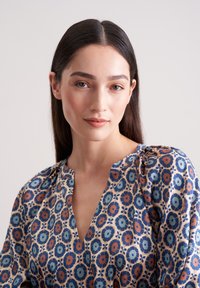 Mehrfarbige Blumenbluse mit V-Ausschnitt, die blaue, orange und grüne Muster auf einem hellen Hintergrund zeigt. Aus glattem Stoff gefertigt.