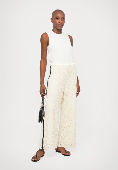 Calças largas de renda creme com riscas pretas nas laterais, combinadas com um top justo branco sem mangas. A modelo segura uma pequena bolsa preta.