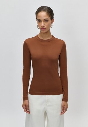 Žena v priliehavom hnedom tričku s dlhými rukávmi a voľných bielych nohaviciach stojaca pred jednoduchým svetlosivým pozadím.