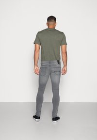 Vaqueros ajustados de color gris con bolsillos traseros, combinados con una camiseta verde de manga corta, vistos desde atrás, sobre un fondo blanco liso.