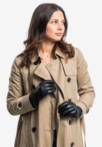 Beige Trenchenmantel mit schwarzen Knöpfen, über einem hellen Oberteil geschichtet. Schwarze Lederhandschuhe mit gesteppten Details und schmaler Passform.