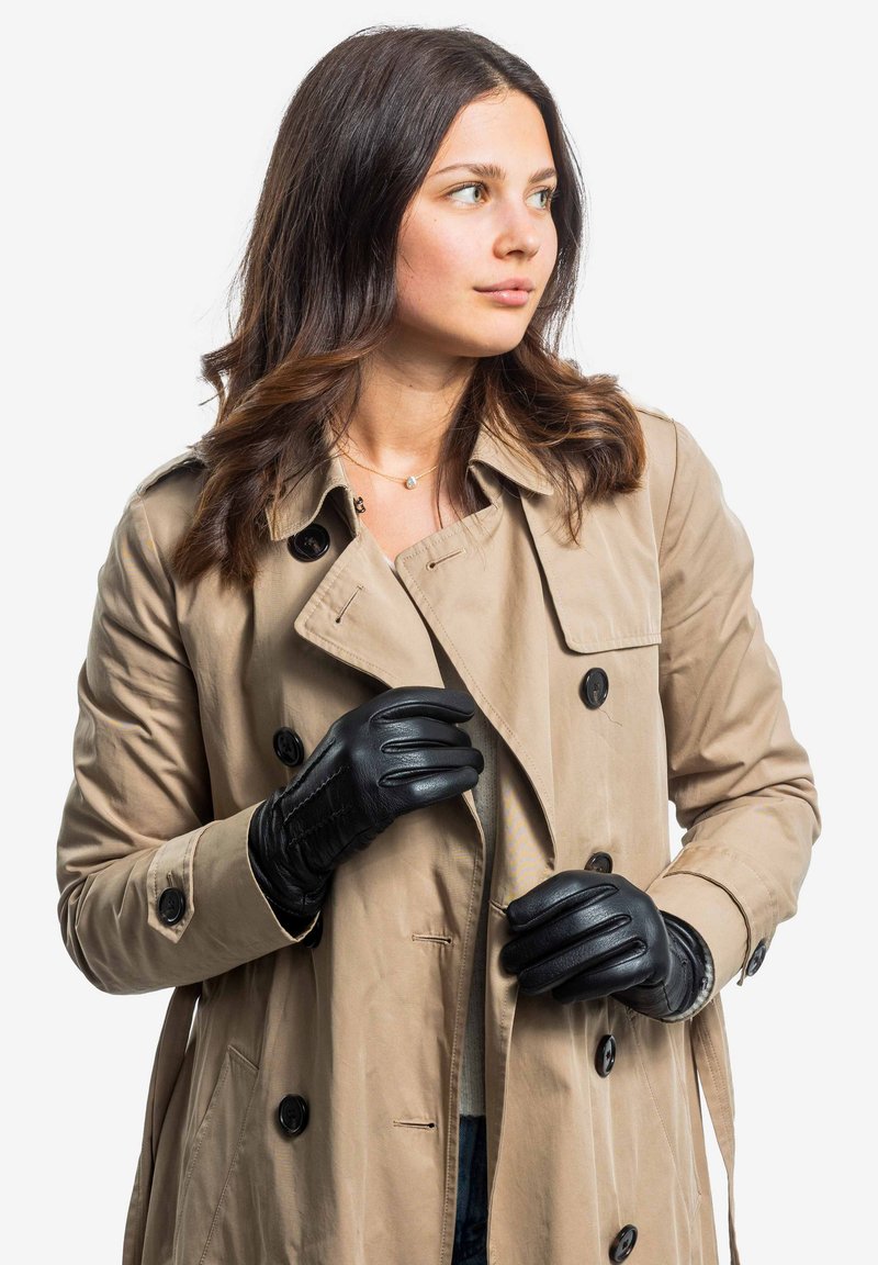 Beige Trenchenmantel mit schwarzen Knöpfen, über einem hellen Oberteil geschichtet. Schwarze Lederhandschuhe mit gesteppten Details und schmaler Passform.