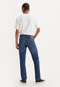 Homme aux cheveux tressés portant un t-shirt blanc, un jean bleu et des baskets noires, debout, une main dans la poche, de dos sur un fond uni.