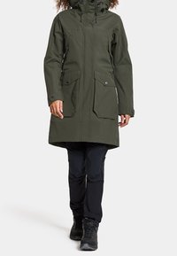 Olivengrüne wasserdichte Parka mit Kapuze, zwei großen Vordertaschen und Knopfverschlüssen. Kombiniert mit schwarzen Hosen und robusten schwarzen Stiefeln.