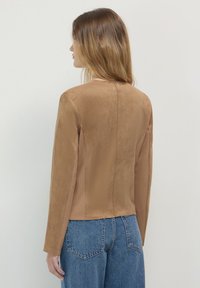 Donna con lunghi capelli castano chiaro, che indossa una giacca di suede beige e jeans blu, vista di spalle su uno sfondo chiaro e semplice.