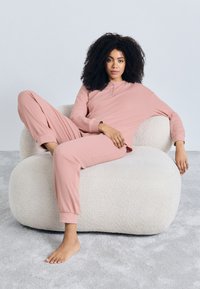 Svetloružový súprava na voľný čas s zapínaním na gombíky a zúženými nohavicami, nosená počas sedenia na textúrovanom, veľkom krémovo bielom kresle.